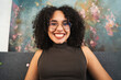 © Studio Marmellata - A young woman with curly hair and glasses smiles warmly while sitting on a couch in front of a colorful abstract painting.