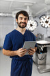 © sofiko14 - A smiling surgeon in scrubs holds a tablet in an operating room, with surgical lights and equipment in the background.