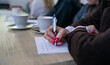© Aga Bak - Close-up of a woman’s hand writing notes during a meeting or workshop. Coffee cups and blurred participants in the background. Concept of learning, education, and creative process.
