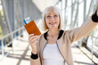 © Anastasiya - Smiling mature woman taking selfie with passport and ticket at airport corridor