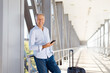 © Anastasiya - Smiling mature man holding smartphone at airport corridor with suitcase beside him