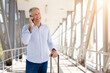 © Anastasiya - Smiling senior man on phone call at airport with suitcase, enjoying communication