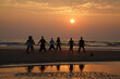 © DragonImages - Silhouettes on unrecognizable people practicing yoga and meditating together at sea shore. Water surface reflecting rays of setting sun. High waves running on beach
