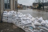 A barrier of white sandbags is piled up alongside a structure to block floodwater from coming in. The scene behind reveals a submerged urban area, highlighting increased water levels following