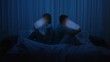 © kinomaster - A couple sits back to back in bed at night using smartphones