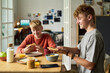 © pressmaster - Two Caucasian teenage boys sitting at table eating breakfast together, smiling and holding toast, smartphone and breakfast foods visible on table, casual home setting