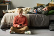 © pressmaster - Caucasian teenage boy sitting on floor using smartphone while Caucasian teenage brother lying on bed holding phone, both engaging with devices in shared bedroom setting
