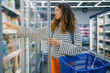 © wifesun - Woman shopping for groceries in supermarket, selecting daily products from refrigerated cooler, holding an empty blue shopping basket, making a healthy food choice
