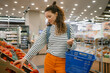 © wifesun - Woman shopping for fresh organic produce, choosing ripe red tomatoes in the supermarket produce section, holding a blue basket, focusing on healthy eating and customer experience