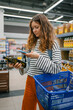 © wifesun - Woman standing in a grocery store, holding an olive oil bottle and a mobile phone, using a shopping list app or researching product details while carrying a blue basket