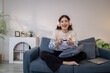 © PHOTO4U - A young woman sits on a sofa wearing headphones while holding a game controller and playing a video game on her laptop. Her cheerful expression shows enjoyment and excitement.