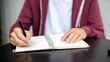 © bongkarn - A man or student holding a ballpoint pen writing in notebook on black table in a cafe or library.