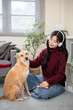 © bongkarn - Pretty asian woman with headphones patting puppy dog while sitting on floor carpet in a living room.