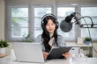 © bongkarn - Woman reading script on clipboard as hosting radio or podcast with laptop and headset at home studio