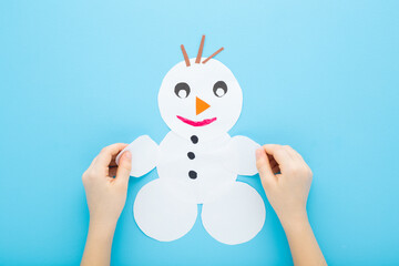 Wall Mural - Little child boy hands holding and showing snowman shape from white paper on light blue table background. Pastel color. Making winter decor. Closeup. Point of view shot. Top down view.