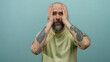 © Krakenimages.com - Man with beard and tattoos appears confused against a blue wall background, touching his head while wearing a pale shirt.
