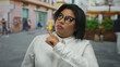 © Krakenimages.com - Latin plus-size woman with glasses gestures thoughtfully on a vibrant city street, showcasing diversity and confidence outdoors.