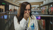 © Krakenimages.com - Hispanic woman doctor with stethoscope laughing in a mall setting, wearing a white coat and standing indoors, blending professional and casual environments.