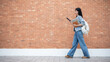 © bongkarn - Asian woman student looking at phone holding laptop and book while walking in front of a brick wall.