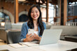 © bongkarn - Smiling asian woman student holding phone over notebook aside laptop while sitting at table in cafe