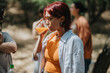 © qunica.com - A young woman with a white shirt over an orange top sips orange juice outdoors, as friends mingle in a sunny park or backyard, capturing a casual, relaxed moment.