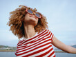 © SHOTPRIME STUDIO - A young woman with curly hair wearing a red striped shirt and bold sunglasses stands outdoors, smiling toward the sky as a gentle breeze lifts her carefree expression.