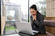 © amnaj - Young Asian businesswoman smiling, celebrating success, working on a laptop during a video call in a modern office setup