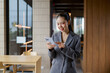 © amnaj - Asian businesswoman standing in a modern office space, smiling and interacting with her mobile device