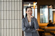 © amnaj - Asian businesswoman in a grey blazer smiling and looking at the camera, posing confidently in a contemporary office setting