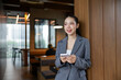 © amnaj - Young Asian businesswoman holding a smartphone, looking away and smiling in a contemporary office workspace