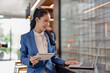 © amnaj - Young Asian businesswoman smiling, working remotely on laptop and holding a financial notebook, standing in a modern office