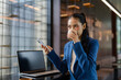 © amnaj - Asian businesswoman sipping coffee while texting on smartphone and working on her laptop in a modern office