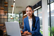 © amnaj - Asian businesswoman working on laptop, confidently smiling in a corporate office setting, symbolizing success and modern business