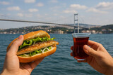 A Person Enjoying a Balık Ekmek on the Waterfront of Istanbul, Holding the Sandwich and Sipping Turkish Tea with the Bosphorus Bridge in the Background