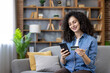 © Liubomir - Smiling woman sitting on a sofa, making an online payment with a credit card and smartphone, enjoying convenient e-commerce and home delivery services