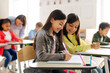 © Home-stock - School girls friends sitting at desks and writing in copybooks, one girl glancing at her friend's test, studying at school classroom interior
