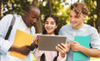 © Prostock-studio - Campus pastime. Happy international teens walking in park, indian lady sharing her tablet, showing new educational app to friends, looking together on device and smiling