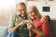 © Prostock-studio - Happy Senior Couple Using Mobile Phone Together, Husband Pointing Finger At Cellphone Showing Something Browsing Internet Sitting On Couch At Home. Mature People And Gadgets