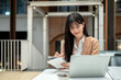 © bongkarn - Asian businesswoman or office worker writing in notebook while looking at laptop on table in a cafe.