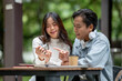 © bongkarn - Young asian couple a man explaining about camera to a woman while sitting at outdoor table in cafe.
