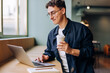 © (JLco) Julia Amaral - Young man working on laptop holding a coffee cup with a smile