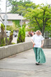 © Masakazu Tokashiki - A woman in a white shirt and green skirt walks down a sidewalk. The scene is set in a residential area with trees and a house in the background. The woman is in a casual