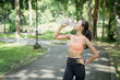 © MINAE - Young woman drinking water from a plastic bottle after a workout in a green park, smiling and refreshed while practicing healthy, active outdoor fitness and hydration