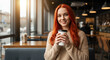 © Madiha - Smiling young woman with vibrant red hair enjoying a warm beverage in a cozy cafe setting, bathed in natural sunlight