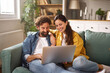 © Stockphotodirectors - Two people are happily interacting while using a laptop on a comfortable couch in a well-decorated living room filled with natural light during the afternoon.