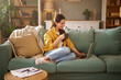 © Stockphotodirectors - Young woman sits comfortably on a green sofa, holding a cup of coffee. She focuses on her laptop, surrounded by books and home decor, creating a warm and inviting atmosphere.