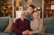 © Stockphotodirectors - A young female home caregiver smiles, posing with a senior couple in their home. The older man and woman are sitting on a green sofa. The caregiver has her arms around them.