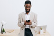 © SHOTPRIME STUDIO - Smiling man with dark skin using smartphone in modern office setting, casual clothing, standing near desk with laptop, stationery, and potted plant, isolated on plain white background, studio shot
