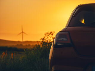 Wall Mural - A red car is parked in a field with a sunset in the background. The car is facing the sun, and the sky is orange and pink. Wind farm turbines in the background. Sightseeing modern technology concept