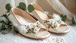 © Sip - Pair of elegant, off-white open-toe shoes with floral embroidery and feather details, set atop lace and near greenery